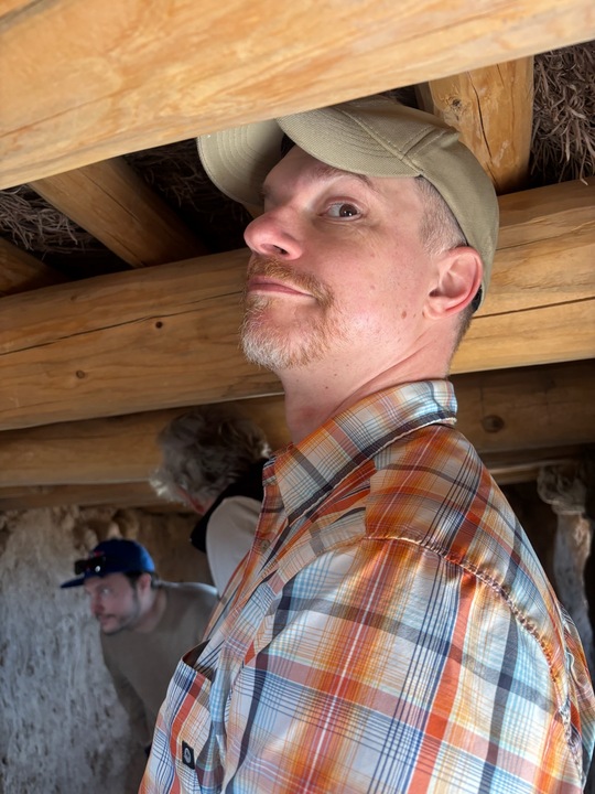 Visitor inside a low-ceiling ancient dwelling at Bandelier National Monument, wearing a cap and plaid shirt.